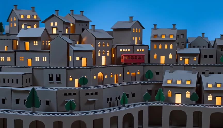 A red paper tram passing cardboard houses as their windows light up at blue hour