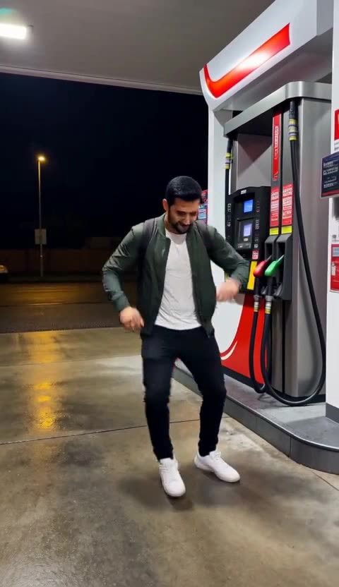 A man in a green jacket dances beside a fuel pump under white canopy lights at night, on wet concrete