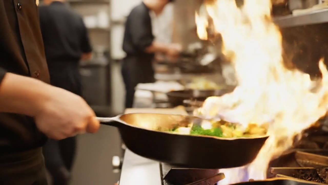 A cook tossing vegetables in a cast-iron pan as flames flare up in a restaurant kitchen